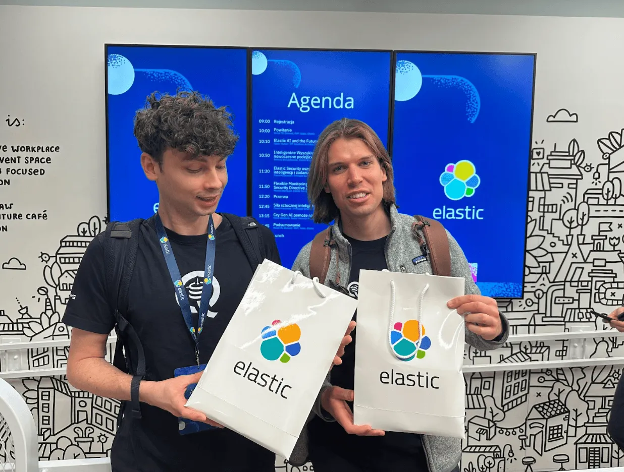 Two men holding Elastic-branded gift bags in front of a conference agenda screen.