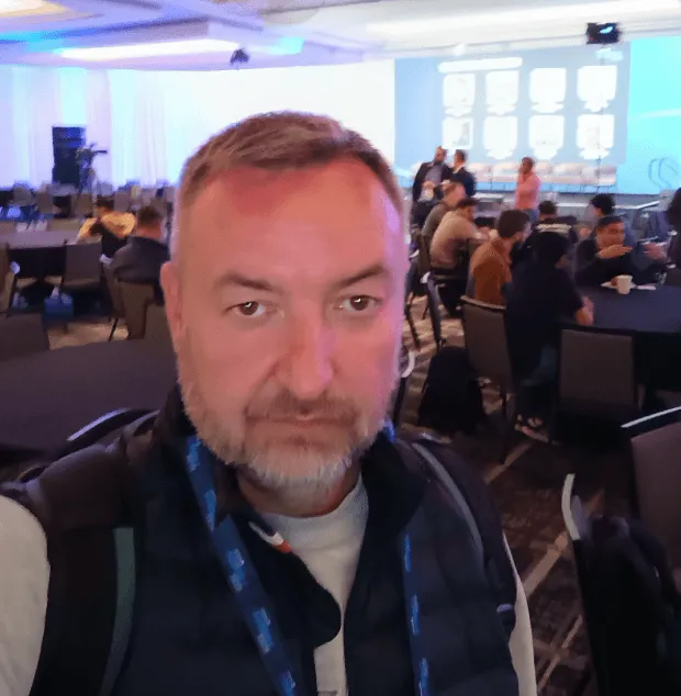 Man taking a selfie at a conference with attendees seated at round tables in the background