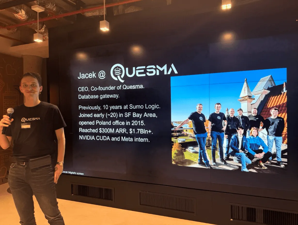 Man holding microphone presenting Quesma talk with team photo and company details on screen