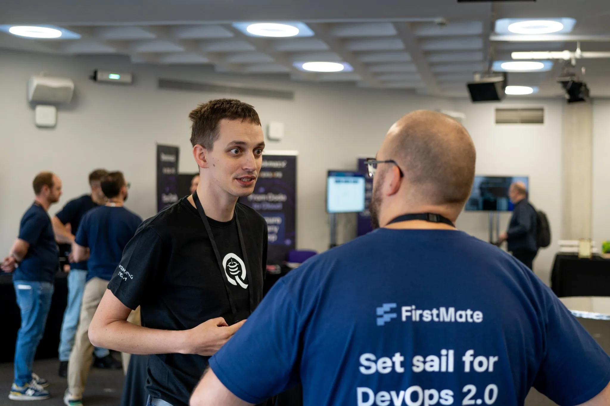 Two people having a conversation at a tech conference, one wearing Quesma shirt and another wearing FirstMate DevOps 2.0 shirt