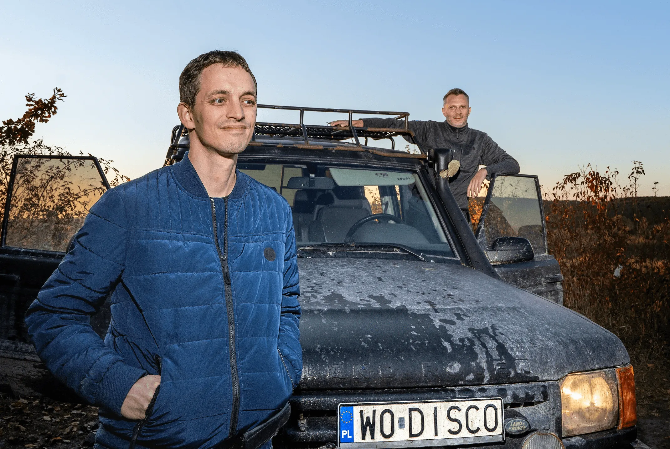 Two men standing by a muddy Land Rover with a custom license plate at sunset.